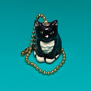 May include: A black and white ceramic cat figurine with a gold-colored ball chain. The cat has green eyes and a pink nose. The figurine is sitting upright. The background is a solid teal color.