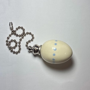 May include: A cream-colored ceramic egg-shaped pull chain handle featuring a vertical row of small light blue dots. The handle is attached to a silver-toned metal ball chain connector and a matching silver-toned metal ball chain.