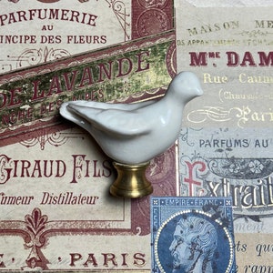 May include: A white ceramic bird figurine with a gold-colored base. The bird is perched on a decorative base, set against a backdrop of vintage French ephemera with text. The figurine is a decorative item.