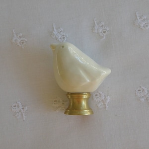 May include: A cream-colored ceramic bird figurine with a glossy finish, mounted on a gold-colored metal base. The bird is perched on a white fabric with embroidered floral details.