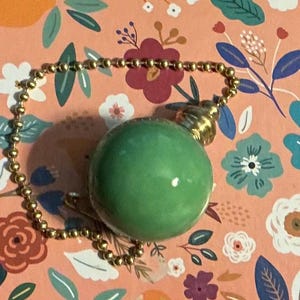 May include: A smooth, spherical green stone pendant attached to a gold-toned metal ball chain. The round green bead features a polished, reflective surface and is connected to a small, ribbed gold-colored metal cap at the top of the sphere.