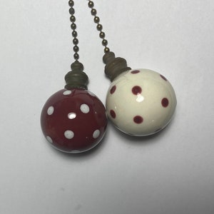 May include: Two decorative ceiling fan pull chains. One is a deep red with white polka dots, the other is white with red polka dots. Both have brass-colored metal accents and ball chains.
