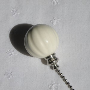 May include: A cream-colored, ribbed light pull with a silver-toned metal base and a ball chain. The light pull is set against a white, textured background, casting a shadow.