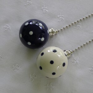 May include: Two decorative light pulls. One is navy blue with white polka dots, the other is cream with navy polka dots. Both have silver-colored metal accents and ball chains. The background is a white, textured fabric.