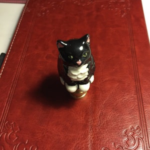 May include: A black and white ceramic cat figurine with green eyes and a pink nose. The cat is perched atop a gold-colored base. The figurine is set on a red leather-bound journal.