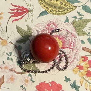 May include: A smooth, spherical red bead attached to a silver-toned metal cap and a matching silver-toned ball chain. The bead has a glossy finish and is connected to the chain through a small metal loop at the top of the spherical component.