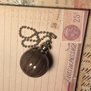 May include: A brown, spherical perfume bottle with a silver-colored metal top and a ball chain. The bottle is resting on a vintage paper background with a postage stamp.