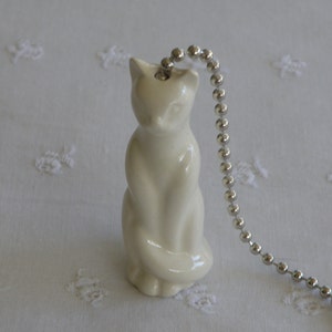 May include: A cream-colored ceramic cat figurine with a silver ball chain attached. The cat is in a seated position with its paws together. The background is a white fabric with a floral pattern.