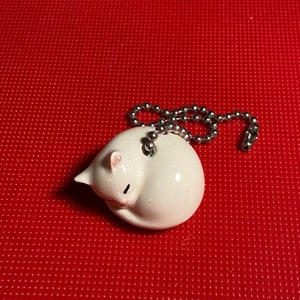 May include: A white ceramic cat figurine curled up in a ball, with pink ears and a black line for a closed eye. It is attached to a silver ball chain. The background is a solid red.