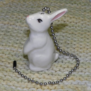 May include: A white ceramic bunny-shaped ceiling fan pull chain. The bunny has pink inner ears and a pink nose. The pull chain is a silver beaded chain with a black connector.