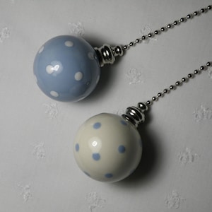 May include: Two decorative light pull chains. One is a pale blue sphere with white polka dots, the other is a cream sphere with blue polka dots. Both have silver-toned metal hardware and ball chains.