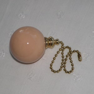 May include: A smooth, spherical light peach-colored pull chain ornament with a polished gold-tone metal cap and a matching gold-tone beaded chain. The round, glossy object features a simple, clean design with a metallic connector at the top.