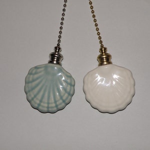 May include: Two ceiling fan pull chains featuring ceramic seashell ornaments. One shell is light blue with darker blue ridges, attached to a silver-toned metal chain. The other is solid white, attached to a gold-toned metal chain with a metal cap.