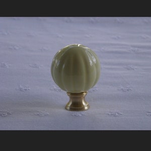 May include: A spherical, cream-colored ceramic knob with vertical fluted ridges. The knob is mounted on a circular, metallic brass-toned base that tapers slightly toward the center, providing a stable foundation for the rounded top piece.