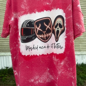 May include: Red t-shirt with a white graphic featuring three masks: a black racing helmet, a neon-outlined mask with an X-shaped eye design, and a Scream mask. The text "Masked men do it better" is printed below the masks.