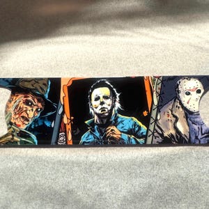 May include: A metal bottle opener featuring horror movie characters. The design includes Freddy Krueger, Michael Myers, and Jason Voorhees. The opener has a floral design on the ends and is primarily black with colorful accents.
