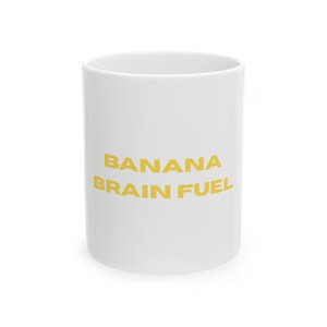 May include: A white ceramic mug with the words "BANANA BRAIN FUEL" printed in yellow. The mug is cylindrical and appears to be empty. The text is centered on the mug.