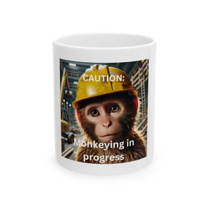 May include: White ceramic mug featuring a monkey wearing a yellow construction helmet. The mug has the text "CAUTION: Monkeying in progress" printed on it. The monkey has brown fur and is set against a construction site background.