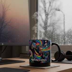 May include: A black coffee mug with a colorful psychedelic design featuring a monkey and the text "Monkey Cosmos." The mug has a black handle and rim, with steam rising from the top. The background includes a computer monitor and a window.