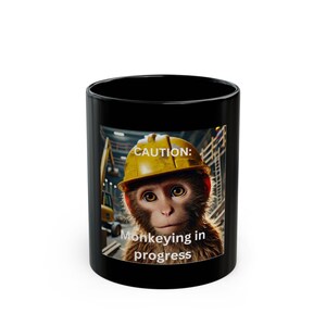 May include: Black ceramic coffee mug featuring a monkey wearing a yellow construction helmet. The mug has the text "CAUTION: Monkeying in progress" printed on it. The background shows construction equipment.