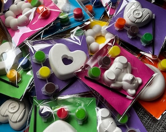Può includere: Una collezione di kit creativi individuali, ognuno contenente una figura in gesso bianco a forma di cuore, orso o ciambella, accompagnata da piccoli vasetti di vernice di vari colori e un pennello nero sottile, sigillati in buste di plastica.