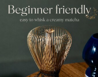 May include: A bamboo matcha whisk with numerous thin tines and a dark, textured handle. The text 'Beginner friendly' and 'easy to whisk a creamy matcha' is visible above the whisk. A dark blue ceramic object is partially visible to the right.