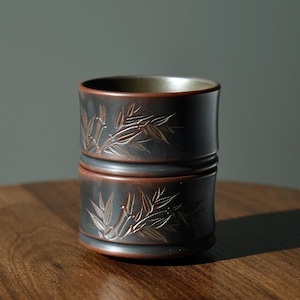 Puede incluir: Una taza de cerámica gris oscuro con un diseño de hojas de bambú grabado en la superficie. El borde y la base tienen un acento marrón rojizo. La taza es cilíndrica y parece hecha a mano.