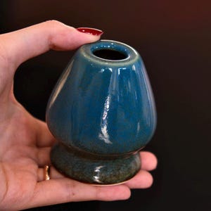 May include: A teal ceramic matcha bowl with a glossy finish. The bowl has a rounded body and a flared opening, and sits on a small, circular base. The bowl is held in a hand against a dark background.