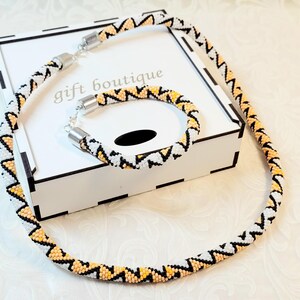 以下が含まれることがあります： 幾何学模様の白とオレンジのビーズネックレスとブレスレットのセット。セットは「ギフトブティック」と書かれた白い箱の上に置かれています。