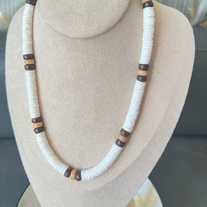 May include: A white and brown beaded necklace with a layered design. The necklace is made of round wooden beads and is displayed on a white mannequin.
