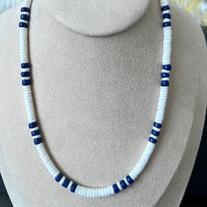 May include: A white and blue beaded necklace with white round beads and dark blue accent beads. The necklace is displayed on a white mannequin.