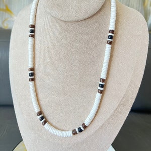 May include: A white, brown, and black beaded necklace made of wood and shell. The necklace is on a white mannequin.