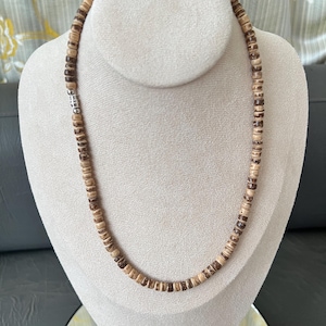May include: A necklace made of small, round, light brown and dark brown wooden beads. The beads are strung together with a silver-colored clasp. The necklace is displayed on a white mannequin.