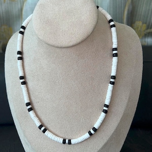 May include: A white beaded necklace with black accents is displayed on a mannequin. The necklace features small, round white beads interspersed with sections of black beads. The necklace is a simple, yet stylish accessory.