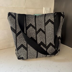 May include: A tote bag featuring a black and white chevron-style arrow pattern with thick black fabric straps. The bag has a rectangular shape and a visible mint green interior lining peeking out from the top opening of the bag.