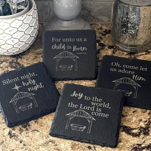 May include: Set of four square black slate coasters with engraved Christmas-themed text and illustrations. Each coaster features a different phrase: "Silent night, holy night," "For unto us a child is born," "Joy to the world," and "Oh, come let us adore Him."
