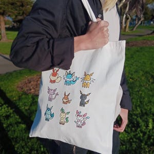 May include: A white tote bag with a colorful cartoon design of various Pokemon characters. The bag has a long strap and is being held by a person.