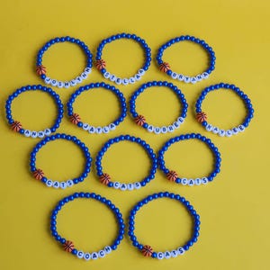 May include: A set of 10 blue beaded bracelets with a basketball charm and white letter beads spelling out names like "Anna", "Jayla", "Aubree", "Kaylee", "Coach", and "Cats".