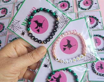 May include: Two bracelets with black and green beads and pink beads. The bracelets spell out "Defy Gravity" and "Glinda". The bracelets are on cards with a witch illustration and a spider web design.