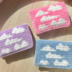 Peut inclure: Trois accessoires rectangulaires en peluche sur le thème des nuages, en rose, violet et bleu. Chacun présente des appliqués de nuages blancs. Les articles sont présentés sur un plateau en bois clair, mettant en valeur une esthétique artisanale.