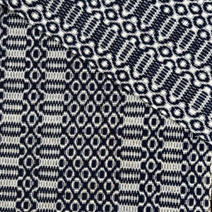 May include: Close-up of a woven textile with a repeating geometric pattern. The design features navy blue and white shapes, including ovals and rectangles, creating a textured surface. The fabric appears to be folded over.