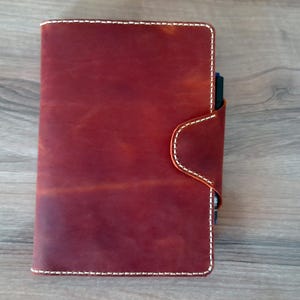Puede incluir: Un diario encuadernado en cuero rojo con costuras blancas y cierre de solapa. El diario está cerrado y sobre una superficie de madera.