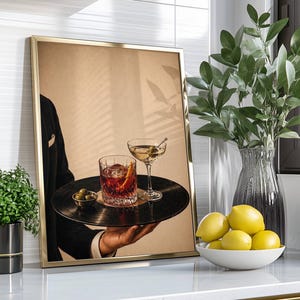 Op de afbeelding: Een ingelijste print met een persoon die een dienblad met twee cocktails en olijven vasthoudt. De dienblad is een vinylplaat. De cocktails zijn een martini en een old fashioned. De lijst is goud.