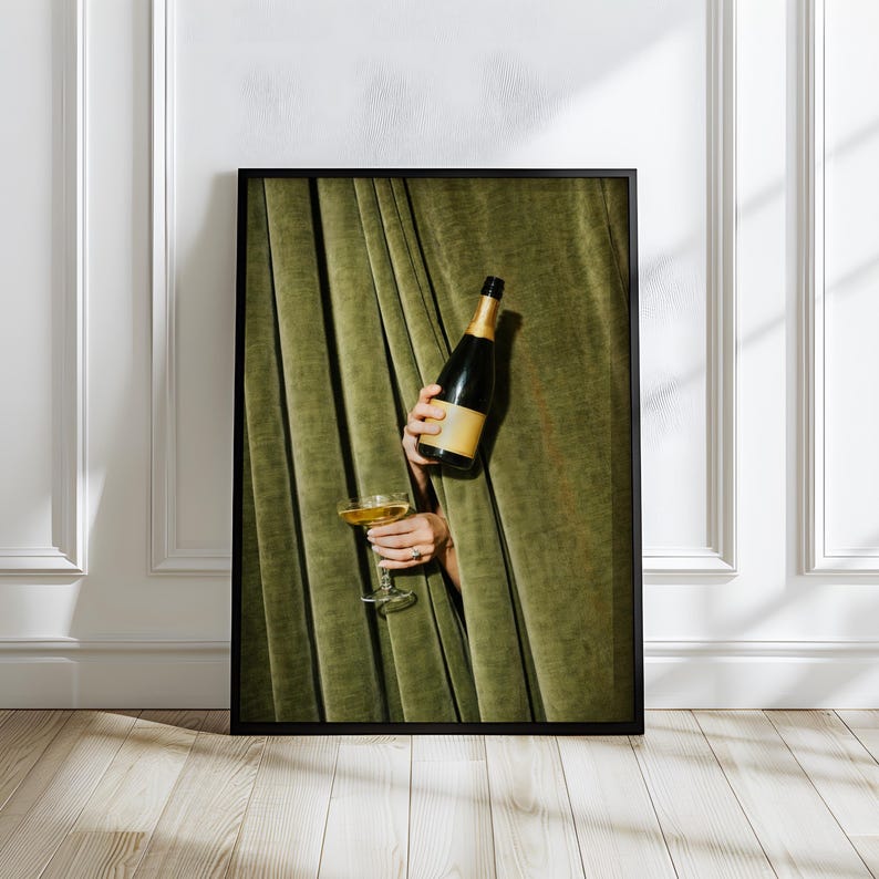 May include: A framed wall art print featuring a dark green velvet curtain backdrop. A hand emerges from the curtain holding a dark glass bottle with a gold foil neck and a clear stemmed glass filled with a golden liquid, all set in a black frame.