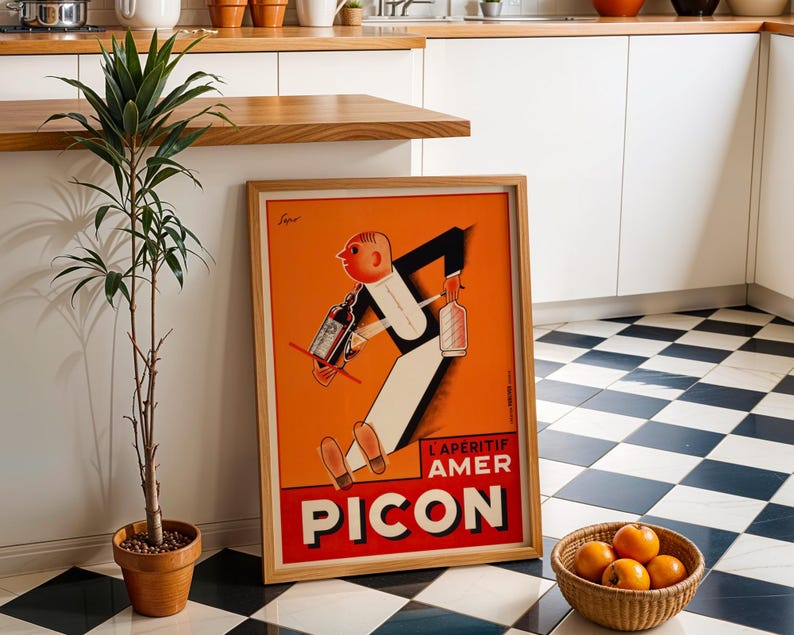 May include: Framed vintage advertisement poster for Picon Aperitif. The poster features a cartoon figure in a suit carrying bottles, set against an orange background. The poster is in a wooden frame and is leaning against a wall next to a potted plant and a basket of oranges.