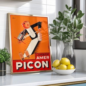 May include: Framed vintage advertisement poster for Picon Amer, featuring a stylized illustration of a waiter carrying bottles. The poster has a gold frame and is set against an orange background with red and white text. A vase of greenery and a bowl of lemons are also visible.