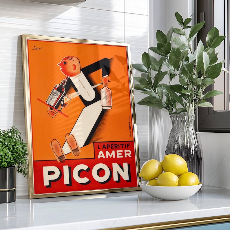 May include: Framed vintage advertisement poster for Picon Amer, featuring a stylized illustration of a waiter carrying bottles. The poster has a gold frame and is set against an orange background with red and white text. A vase of greenery and a bowl of lemons are also visible.