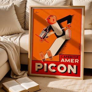 May include: A framed vintage advertisement poster for Pic&oacute;n, an aperitif. The poster features a cartoon figure in a tuxedo carrying bottles and a glass. The background is orange with the words "L'AP&Eacute;RITIF AMER PICON" in red and white.