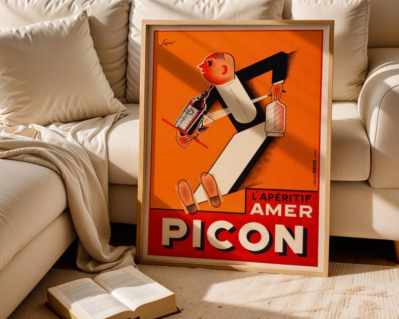 May include: A framed vintage advertisement poster for Pic&oacute;n, an aperitif. The poster features a cartoon figure in a tuxedo carrying bottles and a glass. The background is orange with the words "L'AP&Eacute;RITIF AMER PICON" in red and white.