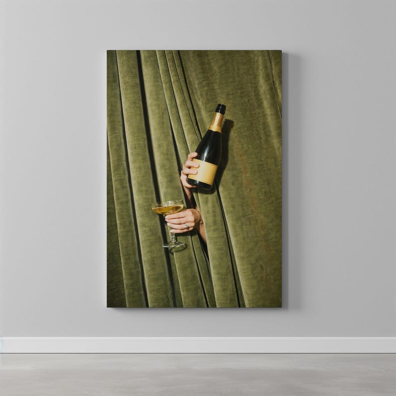 May include: A dark glass champagne bottle with a tan label and a clear glass coupe filled with a golden liquid. The items are held by two hands emerging from behind heavy, pleated olive green velvet curtains that hang in vertical folds.